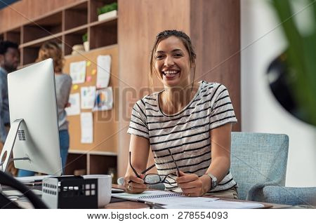 Portrait of smiling young business woman  sitting in a creative agency. Smiling multiethnic woman sitting at her desk in office. Happy casual girl at desktop computer looking at camera.