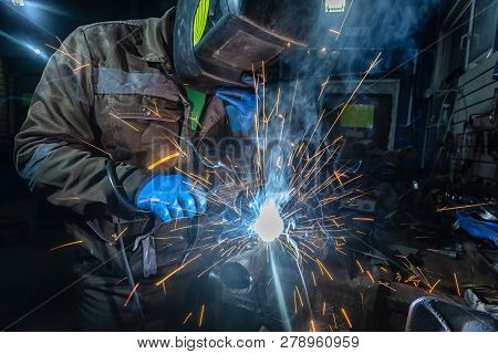 Man Welder In Welding Mask, Building Uniform And Blue Protective Gloves Welds Metal Car Muffler With