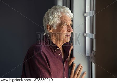 Lonely old man in contemplation near window looking away and thinking. Thoughtful retired man at home looking at view outside window and contemplating. Pensive senior standing and relaxing copy space.