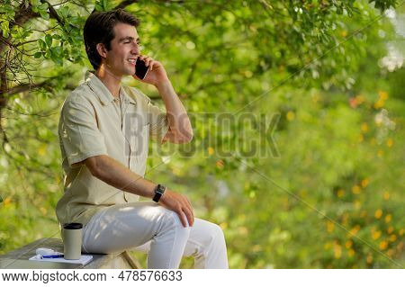 Happy Millennial Man Sitting At Park, Talking On Phone