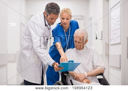 Doctor and nurse with senior patient in wheelchair at hospital corridor talking. Mature doctor showing and discussing medical report with nurse and senior disabled at medical clinic.