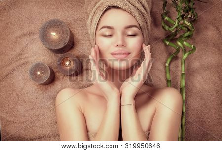 Beautiful spa woman with a towel on her head lying and touching face skin. Skincare. Beauty smiling model girl in spa salon. Skin care, acne treatment, cleansing fresh skin. Renewal cosmetics wellness