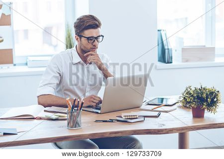 Confident expert at work. Pensive young handsome man using his laptop while sitting at his working place