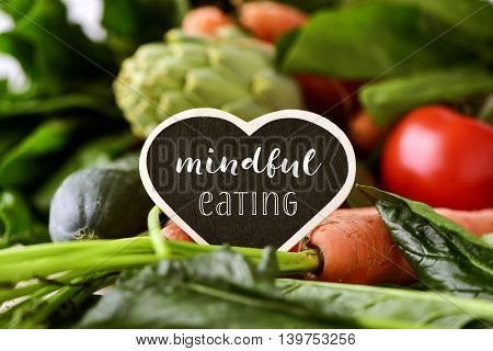 closeup of a heart-shaped chalkboard with the text mindful eating placed on a pile of some different raw vegetables, such as cucumbers, tomatoes, carrots, artichokes, green peppers and zucchinis