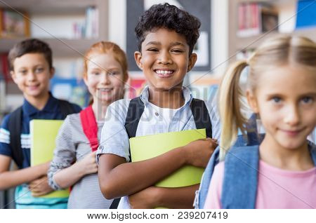 Portrait of a happy multiethnic children holding books and wearing backpack at primary school. Schoolboys and cute girls in a row holding notebook and looking at camera. Elementary child smiling.