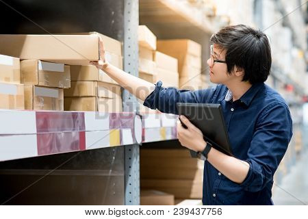 Young Asian Man Doing Stocktaking Of Product In Cardboard Box On Shelves In Warehouse By Using Digit