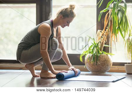 Full length portrait of happy attractive young woman folding blue yoga or fitness mat after working out at home in living room. Healthy life, keep fit concepts. Horizontal image