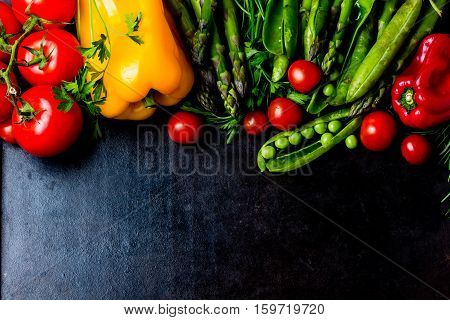 Food background - raw organic vegetables, fresh ingredients for healthily cooking on black background - bell pepper, green pea, asparagus, tomatoes, rosemary. top view. Vegetarian or healthy food