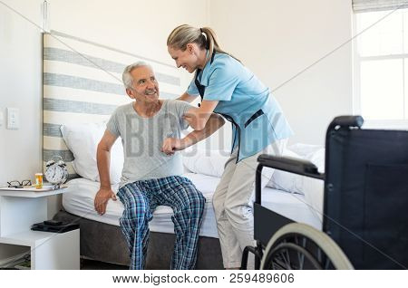 Smiling nurse assisting senior man to get up from bed. Caring nurse supporting patient while getting up from bed and move towards wheelchair at home. Helping elderly disabled man standing up.