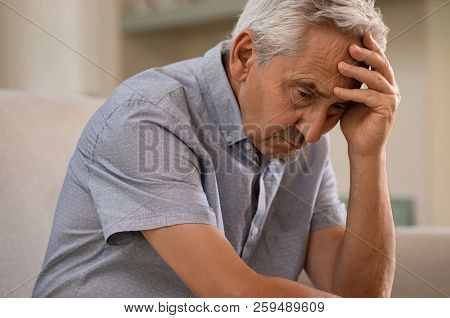 Thoughtful senior man sitting on couch. Depressed sad man sitting with hand on head thinking while looking away. Elderly man suffering from alzheimer.