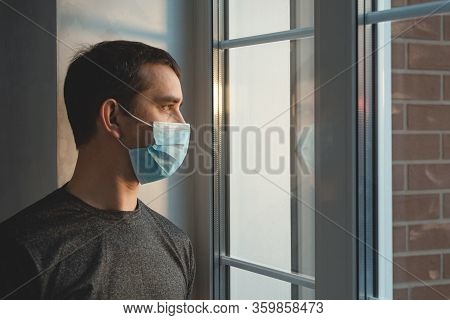 Quarantine Self-isolation. Sad Young Man In A Medical Mask Who Looks Out The Window Through The Wind