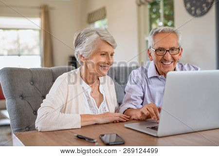 Happy smiling retired couple using laptop at home. Cheerful elderly man and old woman using computer while sitting at table. Smiling pensioner showing woman notebook at home.