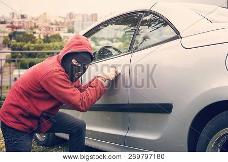 Masked Man Squats And Breaks Someones Car Looking To The Viewer. Thief In Hoodie And Hidden Face Mak