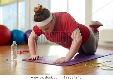 Portrait of young obese woman working out on yoga mat in sunlit fitness studio: performing knee push up exercise with effort to lose weight