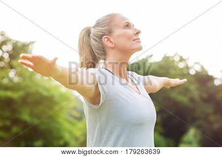 Mature woman doing yoga at park and looking away. Senior blonde woman enjoying nature during a breathing exercise. Portrait of a fitness woman stretching arms and looking away outdoor. 
