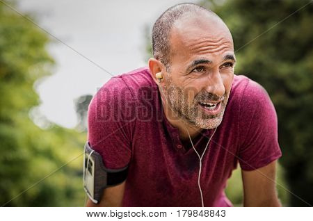 Portrait of athletic mature man after run. Handsome senior man resting after jog at the park on a sunny day. Sweaty multiethnic man listening to music while jogging.