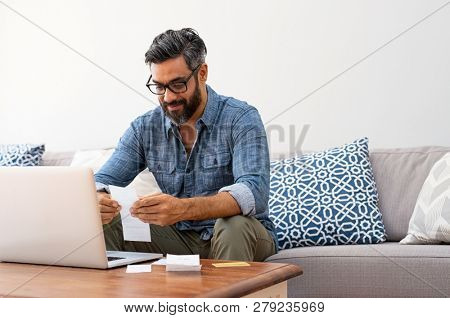 Mature casual man using laptop while looking at invoice. Smiling latin man managing finance with bills and laptop while sitting on couch at home. Hispanic guy reading expenses wearing eyeglasses.