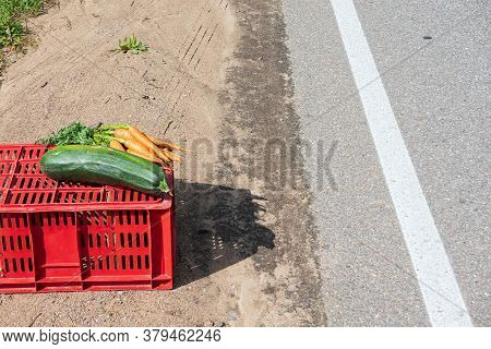 Rustic Vegetables Lie On A Red Plastic Box Near The Edge Of An Asphalt Road. Roadside Trade. There A