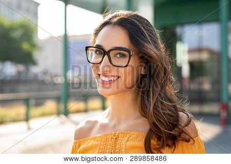 Portrait of carefree young woman smiling and looking at camera with urban background. Cheerful latin girl wearing eyeglasses in the city. Happy brunette woman with long hair and spectacles smiling.