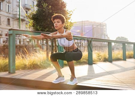 Sporty woman doing squats exercises in the city. Brazilian girl doing sit ups on stairs. Concentrated woman doing warmup squat in urban background.