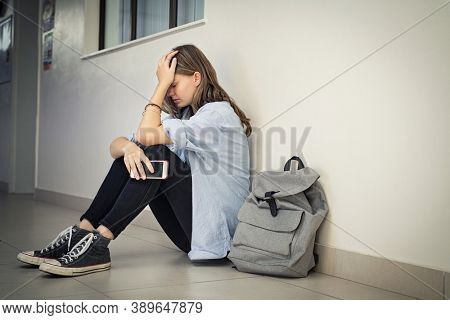 Upset and depressed girl holding smartphone sitting on college campus floor holding head. University sad student suffering from depression sitting on floor at high school. Lonely bullied teen.