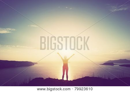 Happy woman with hands up standing on cliff over sea and islands at sunset. Vintage mood, concepts of winner, freedom, happiness etc.