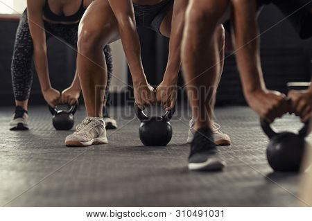 Closeup of young man and fit woman hands lifting kettle bell while squatting at gym. Athlete people doing weight lifting with kettlebell. Group of three young athlete doing fit training.