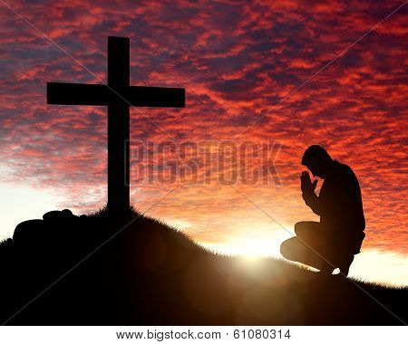 Silhouette of man praying to a cross with heavenly cloudscape sunset concept for religion, worship, love and spirituality