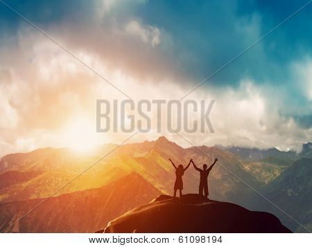 A happy couple standing together on the peak of a mountain with hands raised admiring breathtaking view at sunset