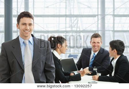 Happy businessman standing in front, businesspeople talking at desk in the background.