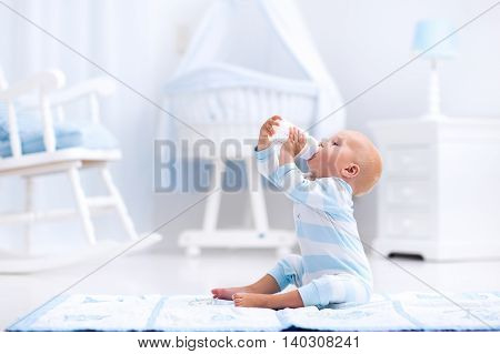 Adorable baby boy playing on a blue floor mat and drinking milk from a bottle in a white sunny nursery with rocking chair and bassinet. Bedroom interior with infant crib. Formula drink for infant.