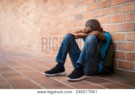 Young boy sitting alone with sad feeling at school. Depressed african child abandoned in a corridor and leaning against brick wall. Bullying, discrimination and racism concept with copy space.