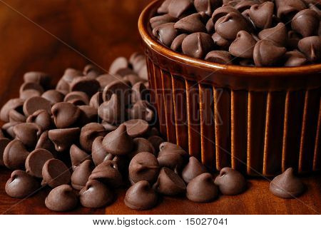 Chocolate chips overflowing from brown ceramic ramekin onto wood table.  Macro with shallow dof.