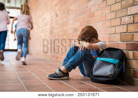 Little boy sitting alone on floor after suffering an act of bullying while children run in the background. Sad young schoolboy sitting on corridor with hands on knees and head between his legs.