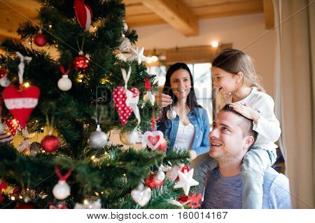 Beautiful young family with little girl decorating Christmas tree at home. Father giving daughter piggyback.