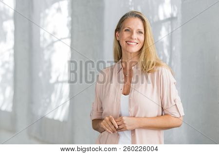 Cheerful mature woman in casual looking away indoor. Successful senior woman smiling and standing confidently. Portrait of happy beautiful lady in a pink shirt with toothy smile standing.