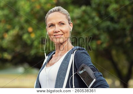 Portrait of a smiling healthy woman with earphones standing in park after running. Proud mature woman looking away after work out. Portrait of satisfied mid woman after fitness exercises.