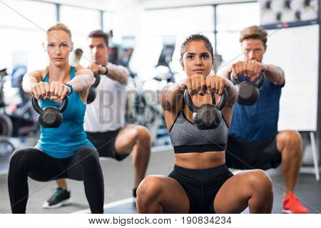 Women and men exercising with kettlebells in gym. Group of young people doing a kettle bell exercise along with squatting. Fitness class and girls training by weights.