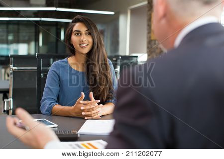 Portrait of young Indian female client or candidate sitting at table, talking to senior male manager and smiling in office. Job interview or consultancy concept