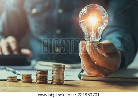 business man hand holding lightbulb with using calculator to calculate and money stack. idea saving energy and accounting finance in office concept