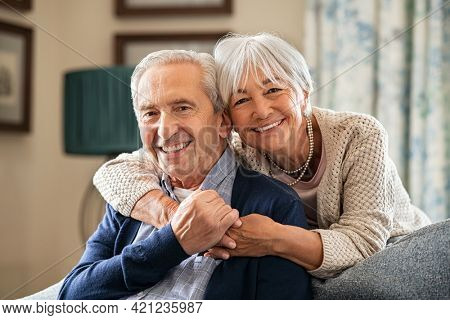 Portrait of romantic senior man with his beautiful wife stay at home. Smiling and caring old woman embracing from behind her retired husband sitting on couch. Cheerful old couple looking at camera.