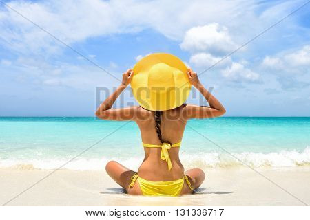 Happy carefree woman relaxing sitting in sand enjoying tropical beach destination. Perfect paradise summer vacation happiness. Back view of bikini girl holding yellow fashion hat on Caribbean holiday.