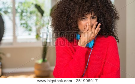 African american woman wearing headphones smelling something stinky and disgusting, intolerable smell, holding breath with fingers on nose. Bad smells concept.