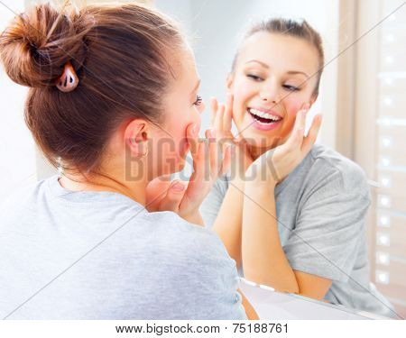 Beauty skin care. Young beautiful teenage girl touching her face before the mirror, enjoying her clean skin. Pretty woman touching her cheek and smiling. Perfect pure skin. Fresh Clean Skin. Skincare