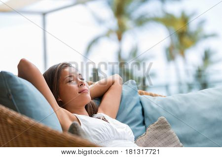 Home lifestyle woman relaxing sleeping on sofa on outdoor patio living room. Happy lady lying down on comfortable pillows taking a nap for wellness and health. Tropical vacation.