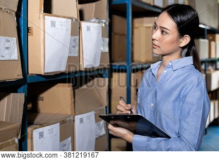 Close up of Asian smart businesswoman working with product inventory and writing on clipboard in distribution warehouse. For business warehouse inventory and logistic concept.