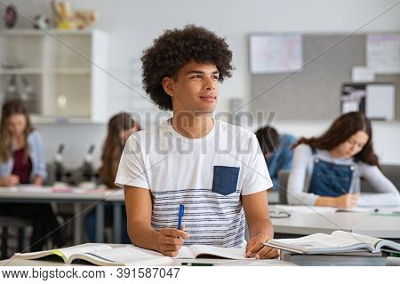 Portrait of contemplative young man during exam at high school. Smiling college student doing homework in class while looking away. Pensive african young man preparing for exam in university classroom