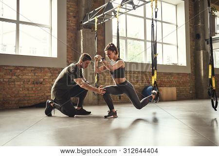 Doing Squat Exercise. Confident Young Personal Trainer Is Showing Slim Athletic Woman How To Do Squa