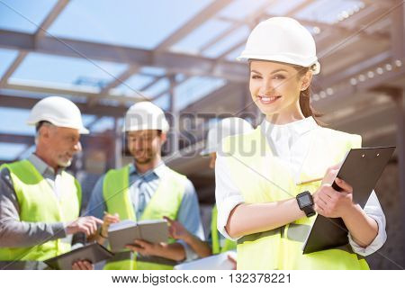 Pretty engineer. A cheerful and content female engineer standing with papers with a group of engineers in a background being in construction site