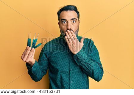 Middle age man with beard and grey hair holding picklock to unlock security door covering mouth with hand, shocked and afraid for mistake. surprised expression 